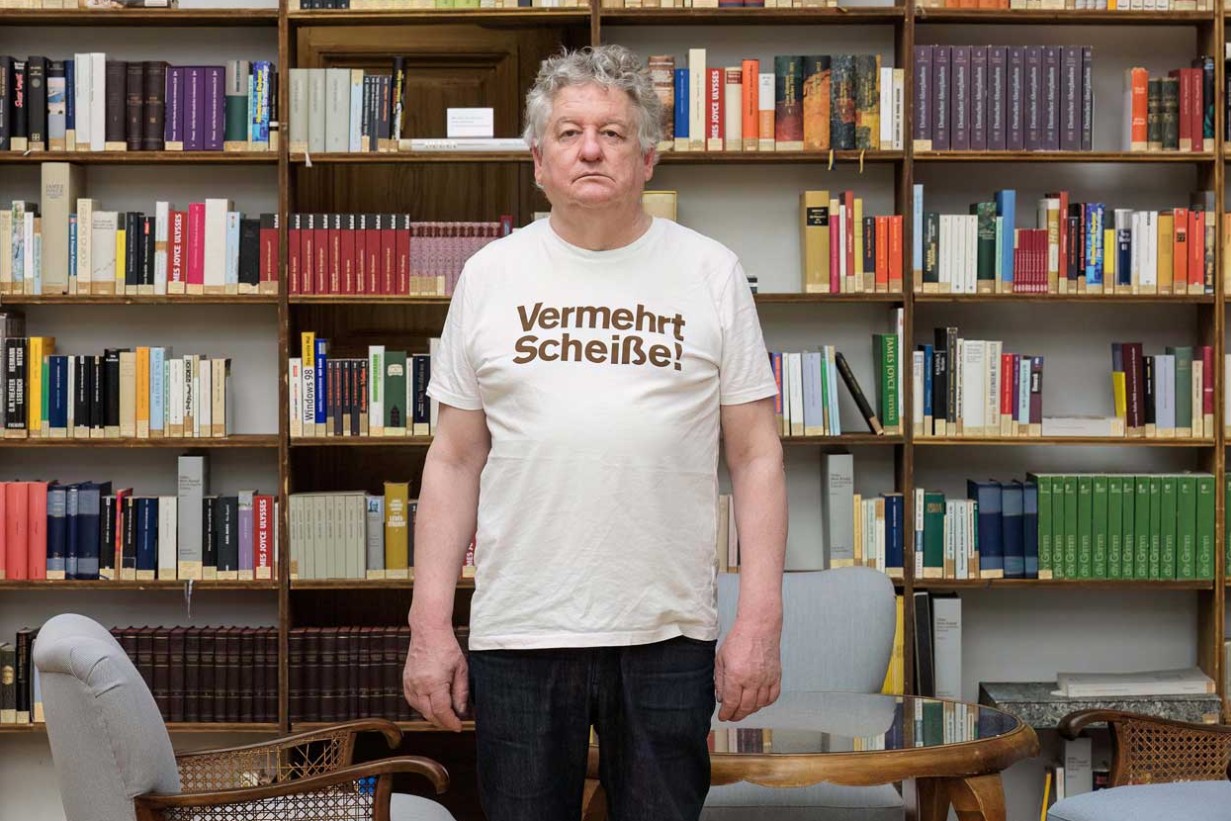 Julius Deutschbauer, ein weißer Mann, steht vor einem Bücherregal. Er trägt ein T-Shirt mit der Aufschrift "Vermehrt Scheiße". 