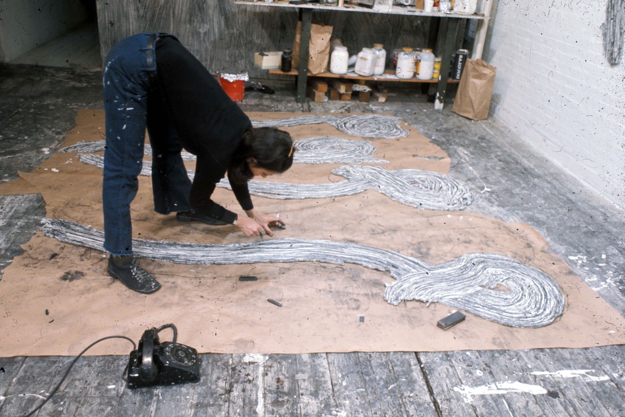A woman bends over to continue working on a sculptural work of art on the floor. In one hand she holds a cigarette.