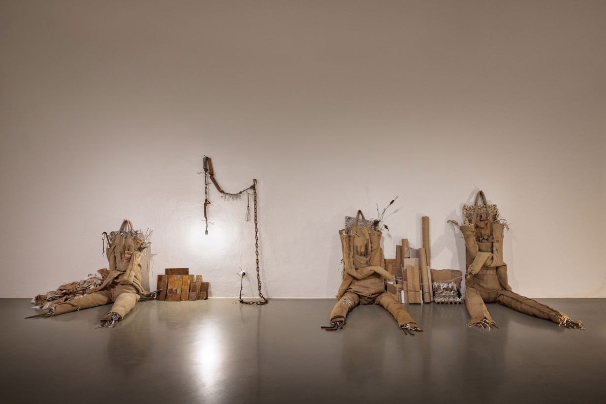 Space with scupltures made of jute bags, leaning against the wall. Accompanying the figures are small sculptures made of scrap metal and electrical wiring, with attached bulbs serving to light the exhibition space.