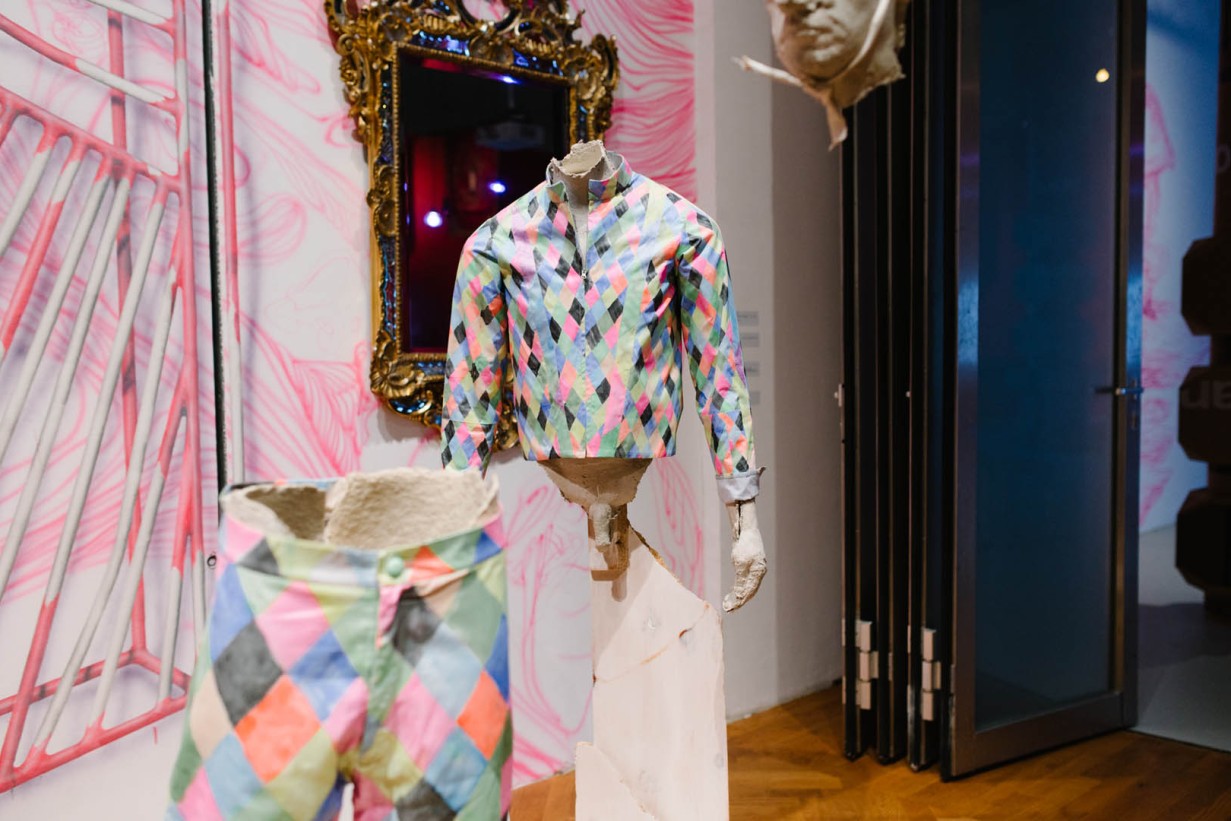 Exhibition room with objects, pink wallpaper with pattern, trousers, figure