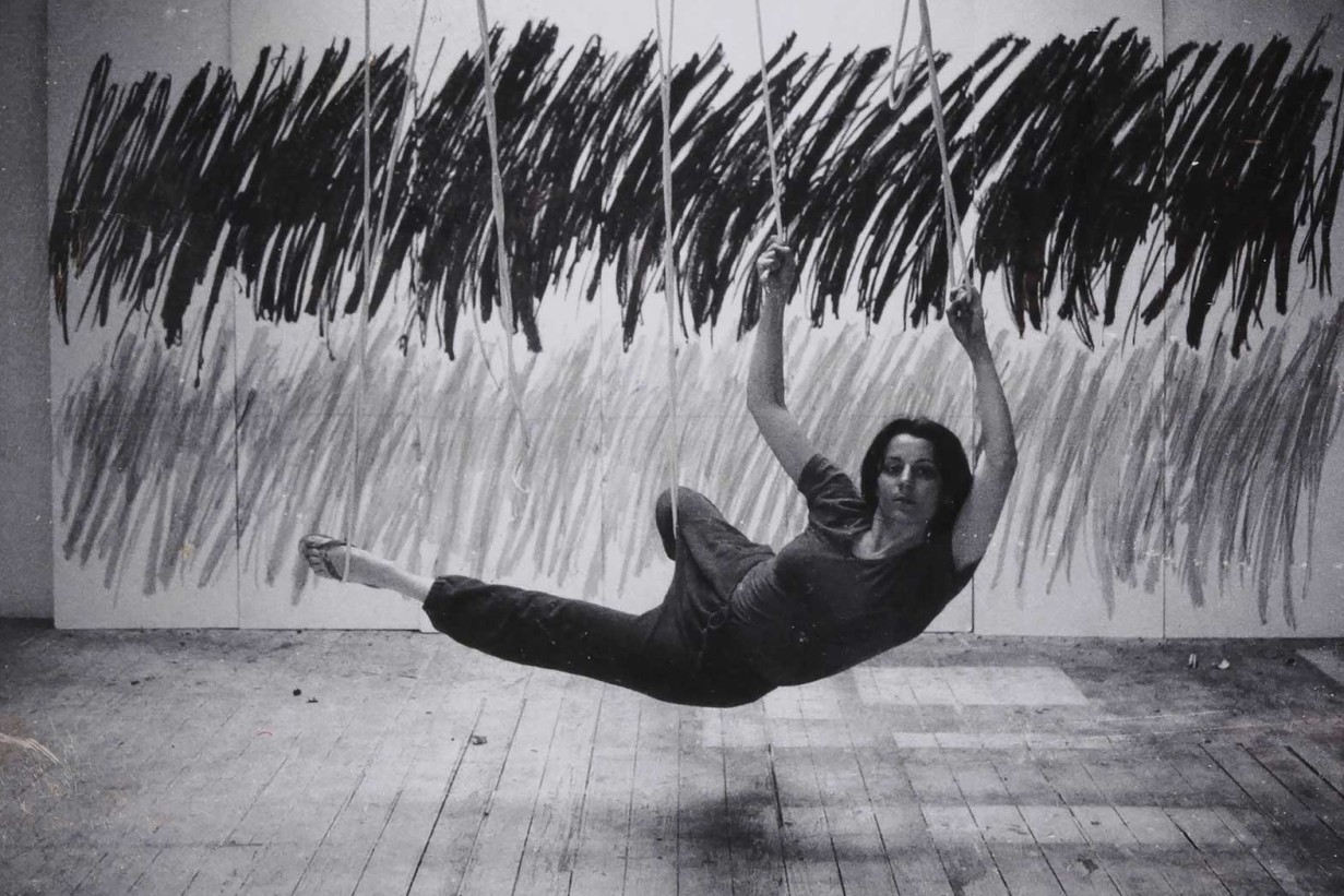 Black and white photograph: a white woman swinging in ropes in front of a work of art. 